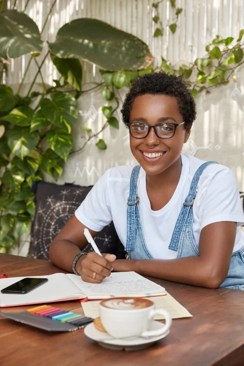photo of cheerful black woman being busy with diary writing or making business plan enjoys hot beve